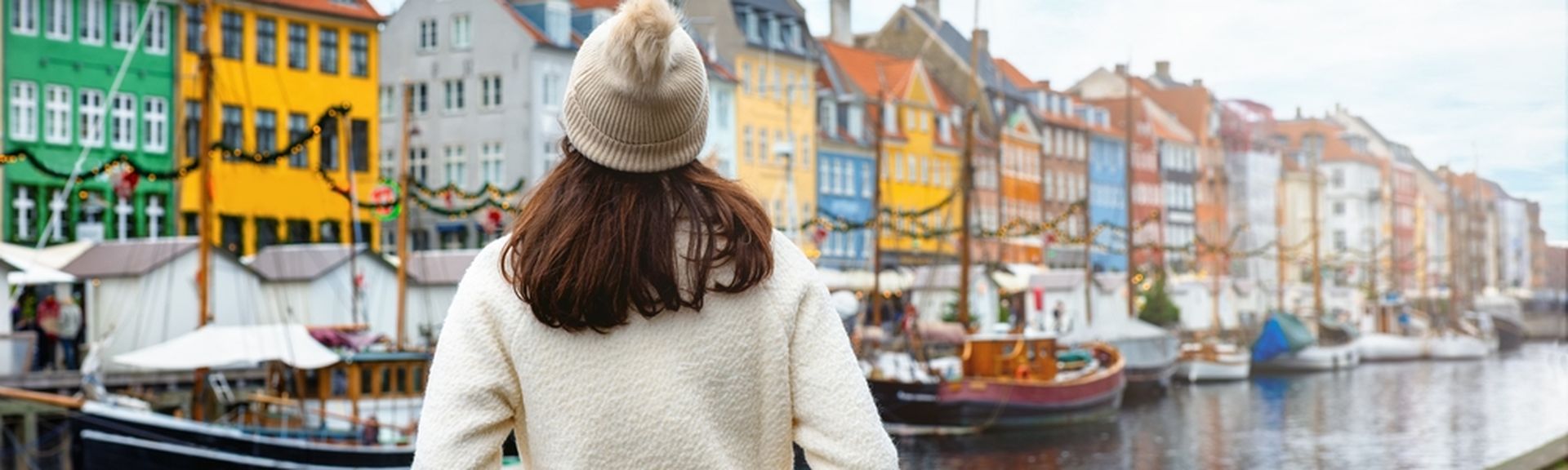 Eine Frau von hinten. Sie trägt eine weiße Jacke und eine Mütze. Sie schaut auf bunte Häuser, das Wasser und einige Boote in Nyhavn in Kopenhagen.