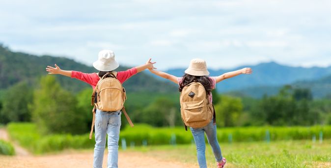 Zwei Mädchen mit Rucksäcken laufen fröhlich einen Weg entlang und heben die Arme in die Luft