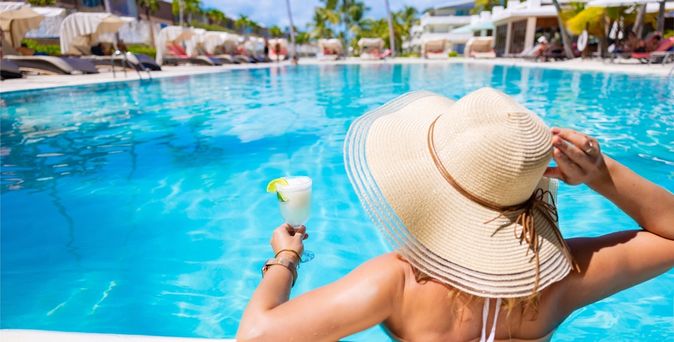 Eine Frau mit Hut von hinten, sie sitzt in einem Pool und hat ein weißes Getränk in der Hand