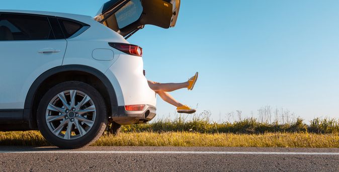 Ein Auto ist zur Hälfte von der Seite zu sehen und steht am Straßenrand an einer Wiese. Der Kofferraum ist auf und man sieht Beine herausragen