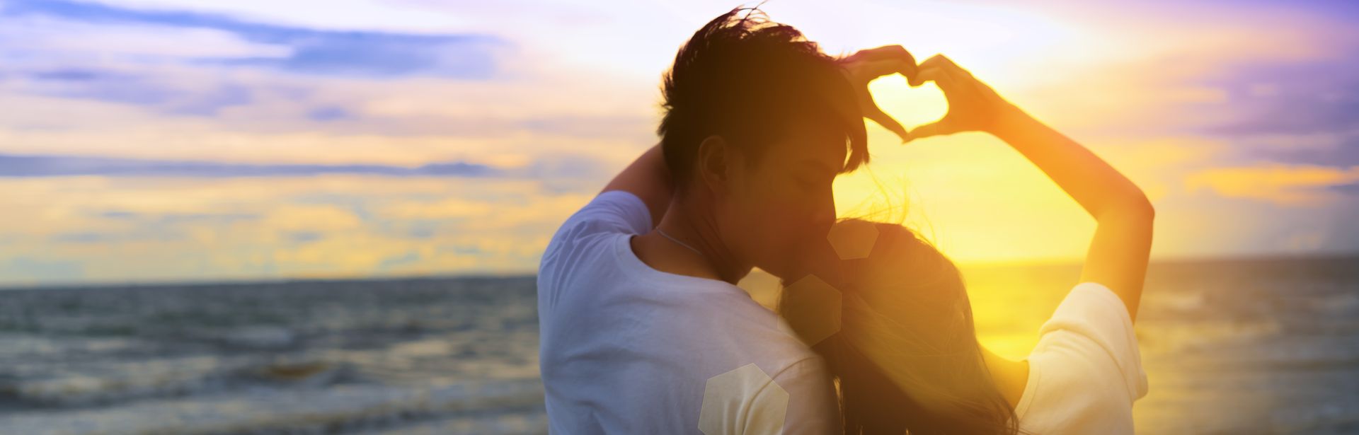 Ein Paar formt mit den Händen gemeinsam ein Herz und schaut bei Sonnenuntergang auf das Meer
