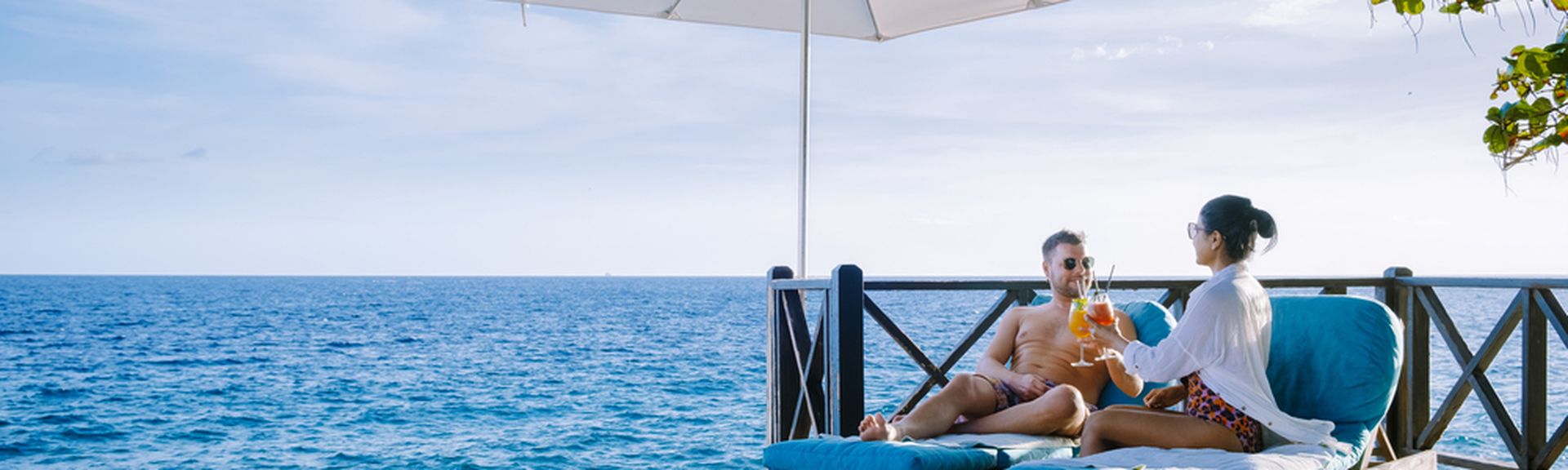 Eine Frau und ein Mann sitzen unter einem Sonnenschirm auf Liegen auf einem Steg und stoßen mit Cocktails an