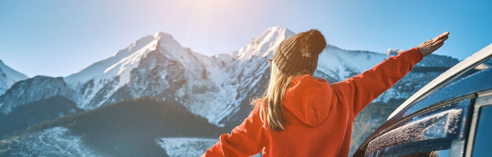 Frau steht an der offenen Tür eines Autos, blickt auf eine schneebedeckte Berglandschaft und streckt die Arme aus