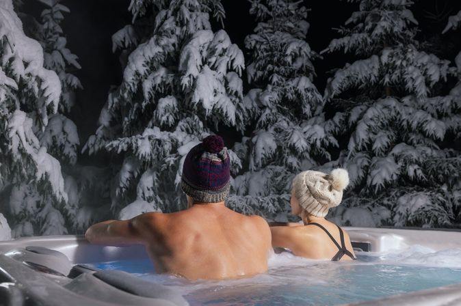 Ein Paar ist trägt Mützen, während sie im Whirlpool sind und nach außen zur Winterlandschaft blicken