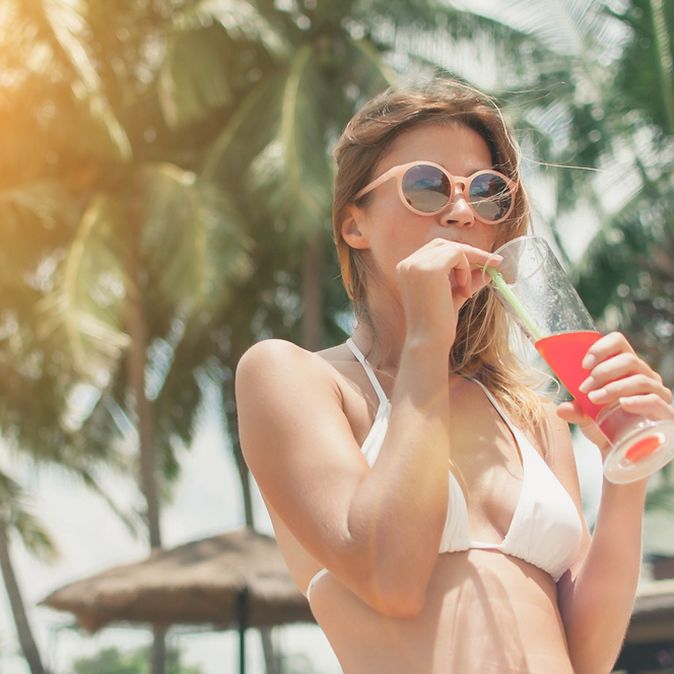 Eine Frau im weißen Bikini und einer Sonnenbrille trinkt mit einem Strohhalm einen roten Cocktail, im Hintergrund sind Palmen zu sehen