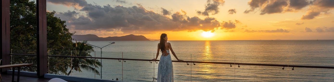 Frau am Geländer einer Terasse schaut auf den Sonnenuntergang über dem Meer