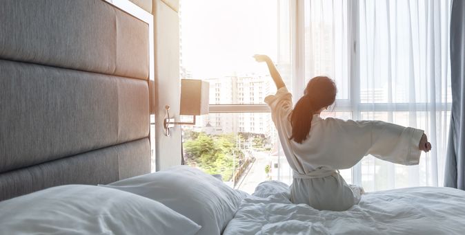 Eine Frau sitzt im Bademantel auf Ihrem Bett und streckt sich 