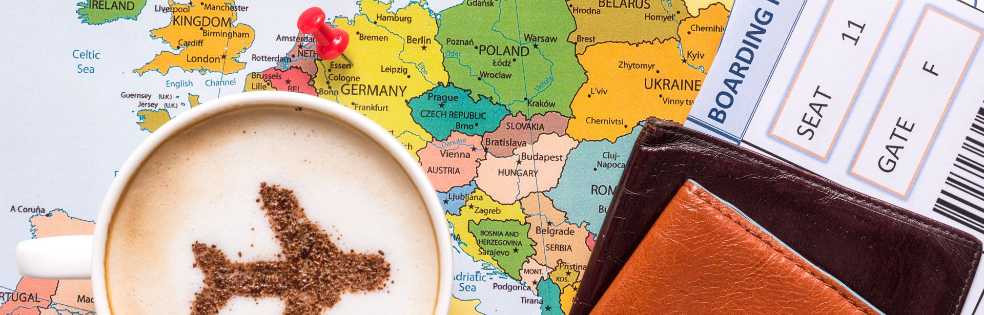 Landkarte mit einem Pin, der in Deutschland gesteckt ist. Auf der Karte steht eine Tasse Kaffee, wo der Schaum die Form einen Flugzeugs ausspart. Ein Flugticket und Lederbörse liegen auch auf der Karte