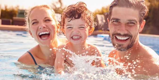 Eltern mit einem Kind spielen im Pool und lachen in die Kamera