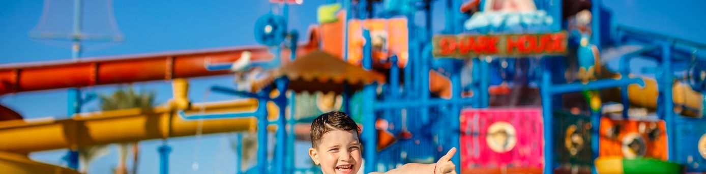 Ein Junge in einer gelben Badehosen steht vor einem großen Aquapark und zeigt einen Daumen nach oben