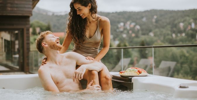 Ein Mann sitzt im Whirlpool und schaut lachen zu einer Frau, die am Rand des Whirlpools über ihm sitzt