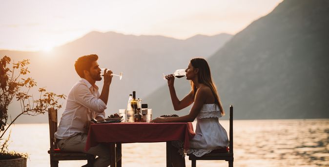 Eine Frau und ein Mann sitzen sich bei Sonnenuntergang an einem Tisch am Wasser gegenüber und trinken Wein