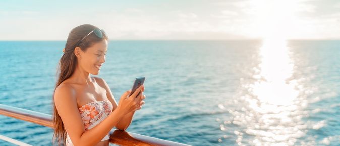 Eine Frau steht lächelnd an der Reling eines Kreuzfahrtschiffes und schaut auf ihr Smartphone