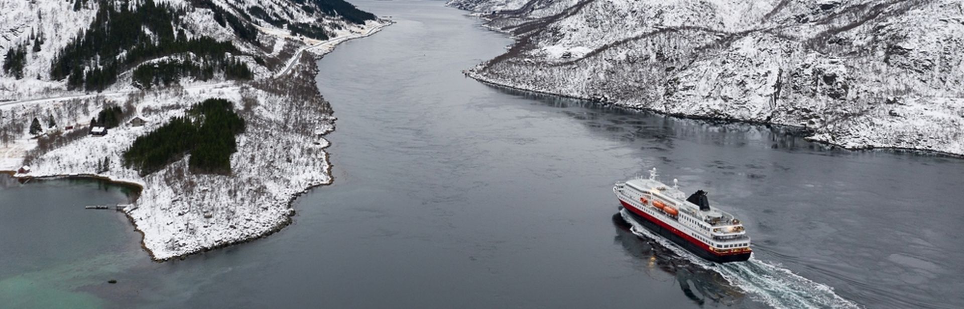 Ein Kreuzfahrtschiff fährt durch einen Fjord, umgeben von schneebedeckten Bergen und winterlicher Landschaft.