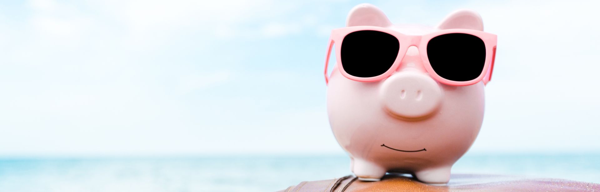 Ein rosanes Sparschwein mit Sonnenbrille sitzt auf einem Koffer am Strand