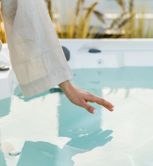 Eine Frau sitzt im Bademantel am Rand eines Whirlpools und hält Ihre Hand über die Wasseroberfläche