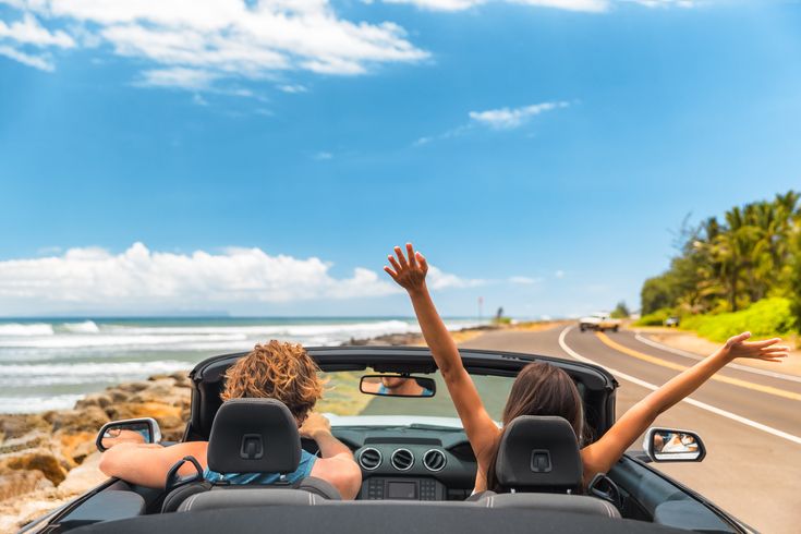 Zwei Personen im Auto mit offenem Verdeck, im Hintergrund kann man das Meer sehen