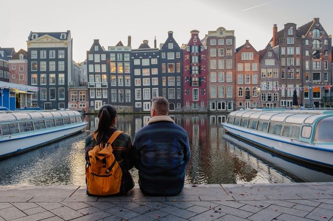 Zwei Personen sitzen in Amsterdam am Wasser und schauen gemeinsam auf die gegenüberliegenden bunten Häuser und die Boote