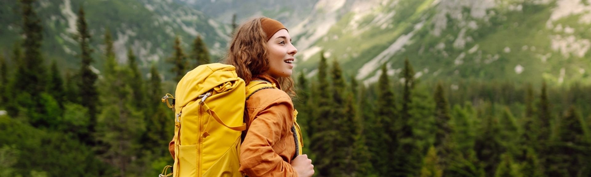 Eine Frau mit gelbem Wanderrucksack steht auf einem Berg und schaut begeistert in die Ferne