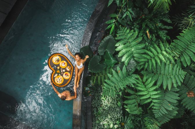 Ein Paar von oben, eine Frau sitzt am Rand eines Pools, ein Mann steht im Pool, zwischen ihnen schwimmt ein Herzförmiges Tablett mit Essen