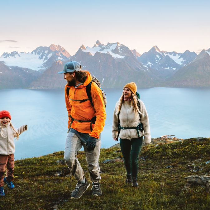 Eine Familie wandert in Winterkleidung an einem Gewässer, im Hintergrund sind verschwommen Berge 