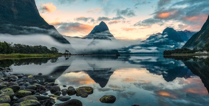 Runde Steine, die in einen See führen im Hintergrund sind von Nebel umzogene Berge zu sehen, die sich im Wasser spiegeln