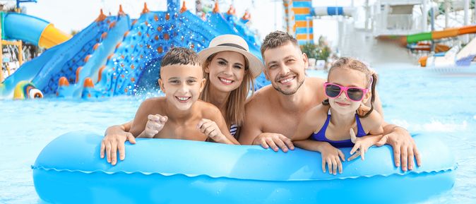 Eine Familie auf einem blauen Pool Floatie lächelt in die Kamera, im Hintergrund ist ein Aquapark