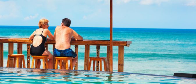 Eine Frau und ein Mann in Badekleidung sitzen an einer Holztheke auf Holzstühlen vor einem Pool und schauen auf das Meer