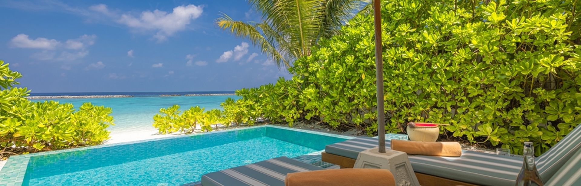 Zwei Liegen unter einem Sonnenschirm, davor ein kleiner Pool und der Strand 