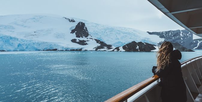 Eine Frau steht mit wehenden Haaren an der Reling eines Kreuzfahrtschiffes und schaut auf schneebedeckte Berge