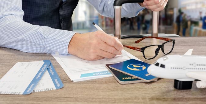 Eine Person sitzt vor einem Tisch und schreibt auf einem Blatt Papier, in der anderen Hand hält die den Griff eines Koffers fest. Auf dem Tisch liegen Flugtickets, eine Brille und zwei Reisepässe