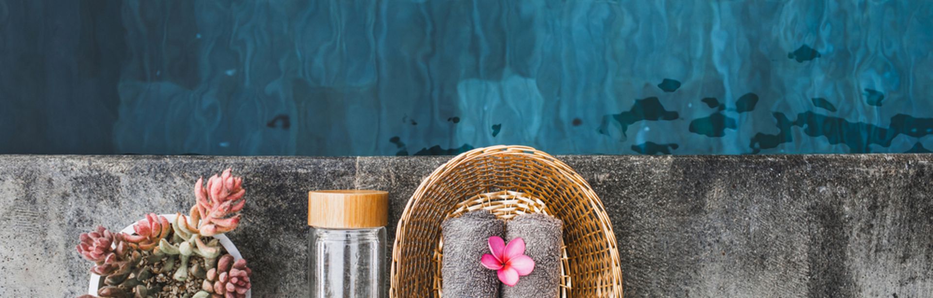 Ein Teil eines Pools aus der Vogelperspektive, ein Korb mit Handtüchern, eine Glasflasche und eine Pflanze im Topf stehen am Rand