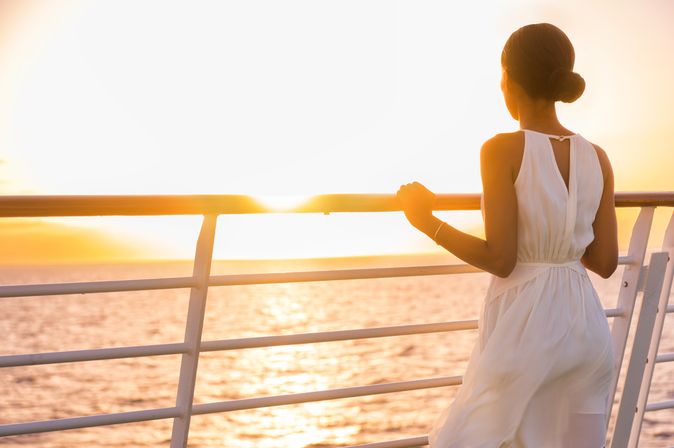 Eine Frau steht an der Reling eines Kreuzfahrtschiffes und schaut in den Sonnenuntergang