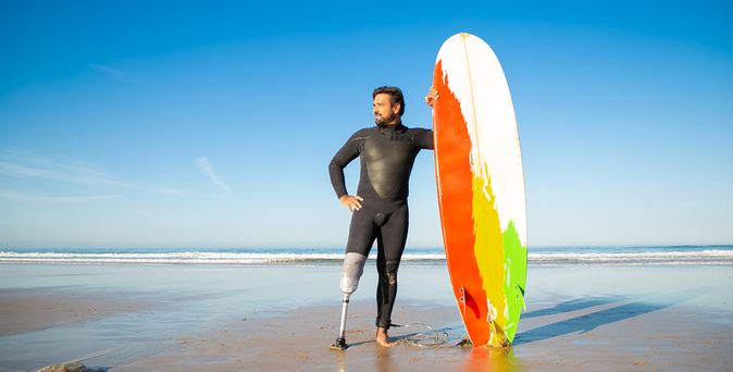 Ein Mann mit einer Beinprothese am linken Bein steht in einem Neoprenanzug am Strand und hält ein buntes Surfboard