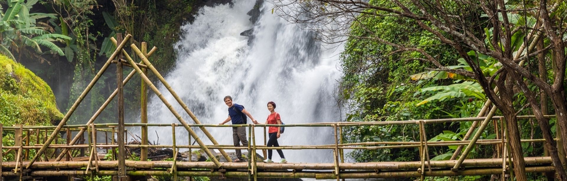 Eine Frau und ein Mann laufen Hand in Hand über eine Holzbrücke über einem Fluss, im Hintergrund ist ein großer Wasserfall