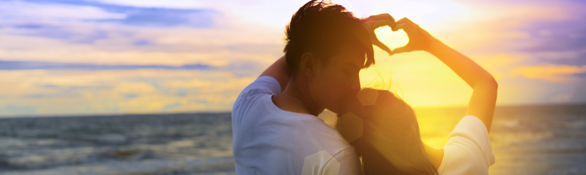 Ein Paar formt mit den Händen gemeinsam ein Herz und schaut bei Sonnenuntergang auf das Meer