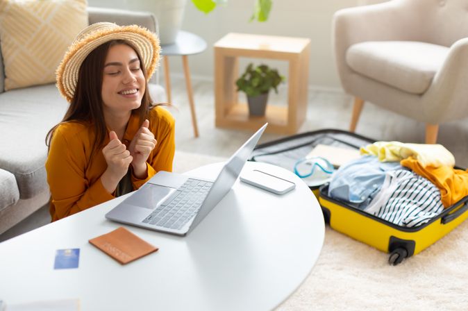 Eine Frau sitzt glücklich vor ihrem Laptop, auf dem Tisch liegen Reisepass und Kreditkarte, im Hintergrund liegt ein gepackter Koffer