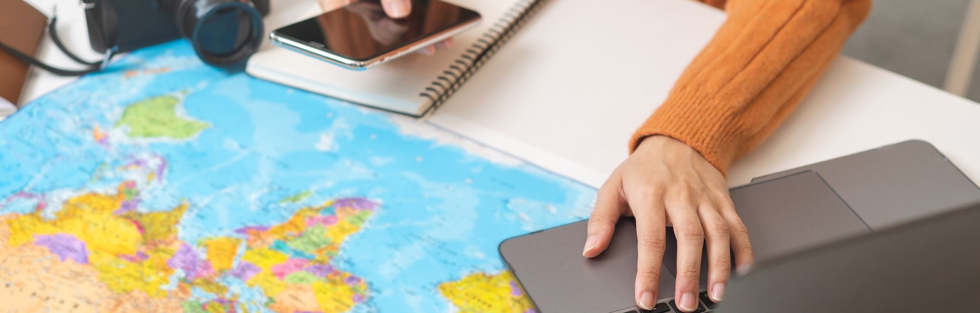 Ein Schreibtisch mit einer Weltkarte als Unterlage, einem Sonnenhut und einer Kamera, eine Person bedient mit der einen Hand ein Handy und mit der anderen einen Laptop