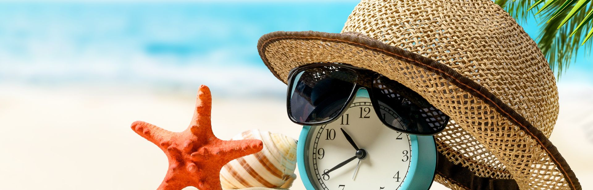 Ein Wecker mit Sonnenhut und einer Sonnenbrille liegen am Strand; Daneben befinden sich Muscheln und ein Seestern