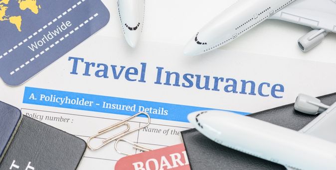 In der Mitte des Bildes steht "Travel Insurance" darum liegen mehrere Büroklammern, Reisepässe und mehrere kleine Flugzeuge