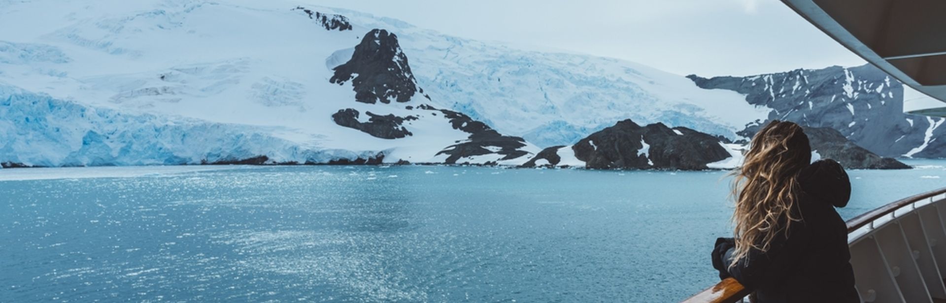 Eine Frau steht mit wehenden Haaren an der Reling eines Kreuzfahrtschiffes und schaut auf schneebedeckte Berge