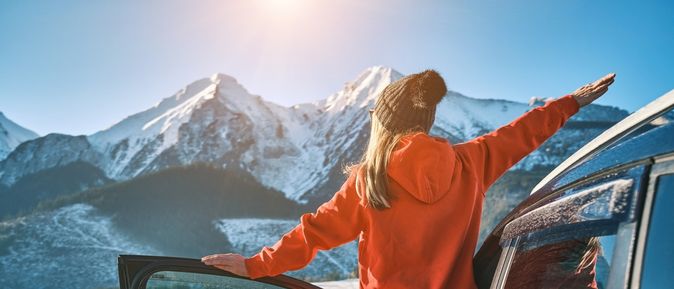 Frau steht an der offenen Tür eines Autos, blickt auf eine schneebedeckte Berglandschaft und streckt die Arme aus