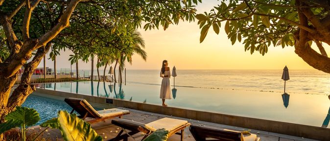 Frau steht bei Sonnenuntergang vor einem Pool der direkt vor einem Meer liegt