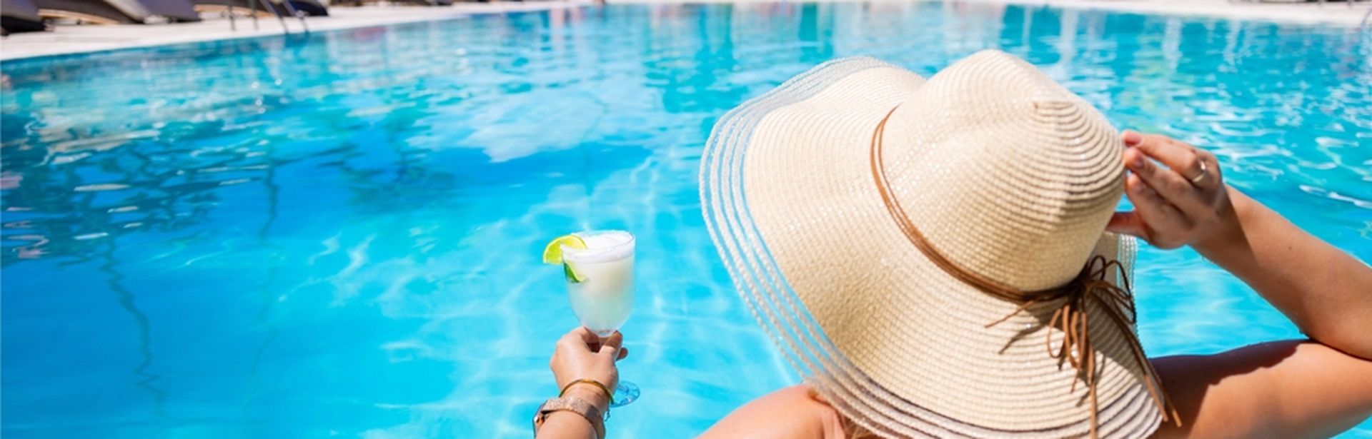Eine Frau mit Hut von hinten, sie sitzt in einem Pool und hat ein weißes Getränk in der Hand