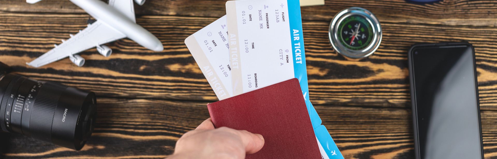 Auf einem Holztisch liegen ein Handy, ein Miniaturflugzeug, eine Kamera, ein Kompass, Geld und ein Globus. Eine Hand hält einen Reisepass und zwei Flugtickets