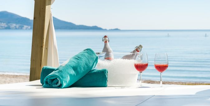 Zwei aufgerollte Handtücher liegen neben zwei Gläsern und ein Getränkekühler mit Eis und zwei Flaschen auf einer Decke am Strand