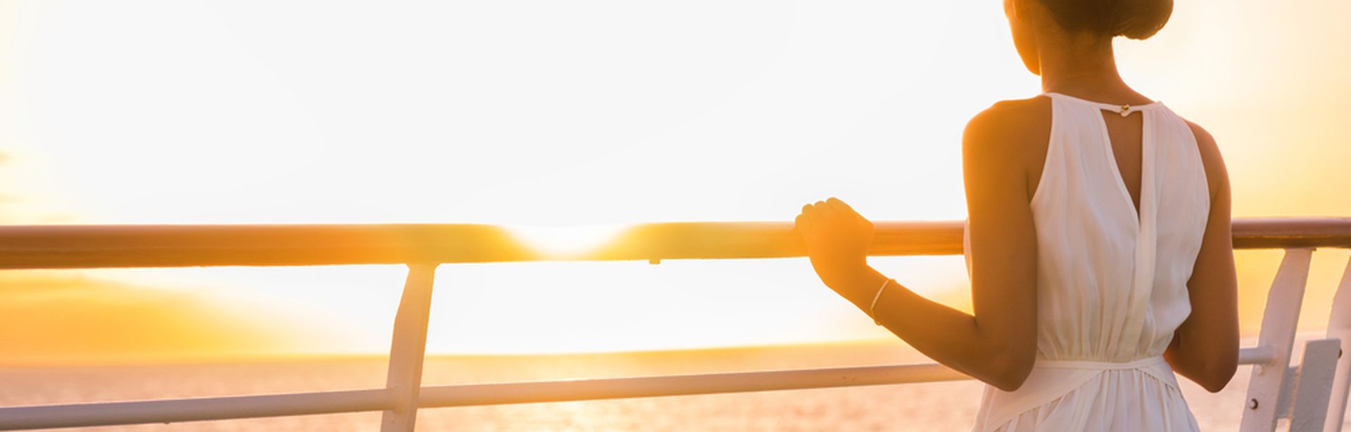 Eine Frau steht an der Reling eines Kreuzfahrtschiffes und schaut in den Sonnenuntergang