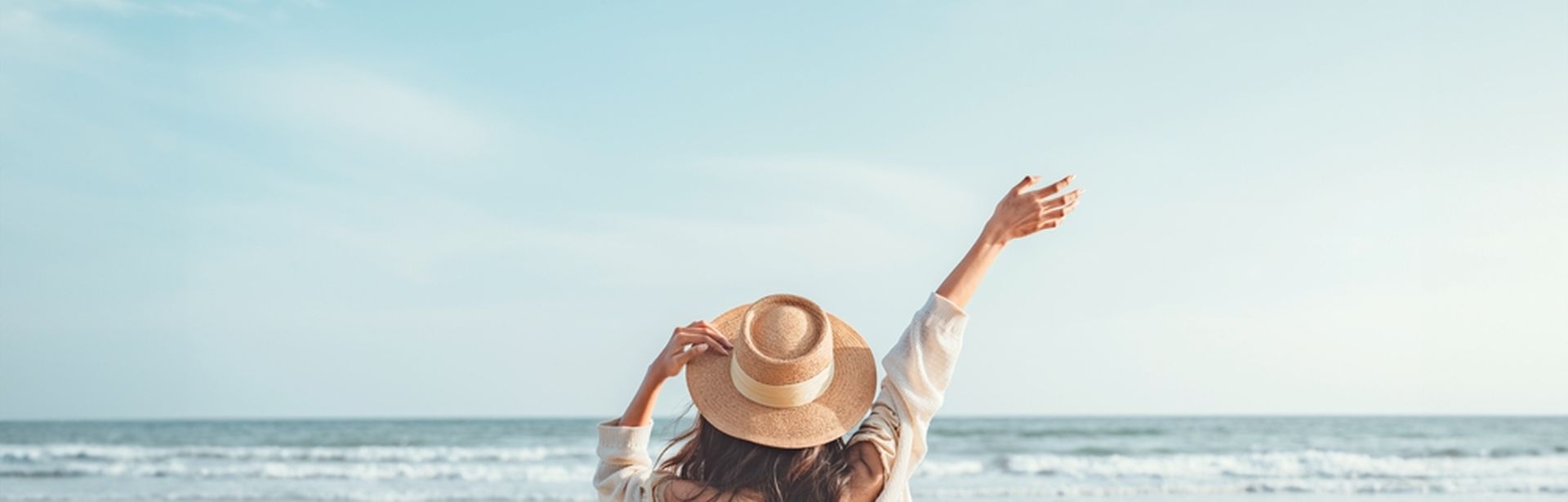 Eine Frau mit Hut von hinten, Sie schaut vom Strand aus auf das Meer und hebt den Arm 