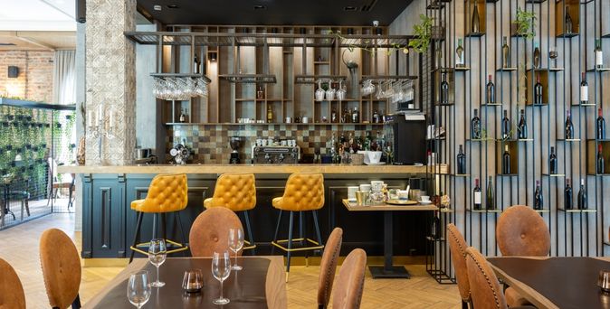 Eine Modern eingerichtete Bar mit gelben Barhockern und einer Wand mit Weinflaschen, im Vordergrund stehen mehrere mit Gläsern gedeckte Tische