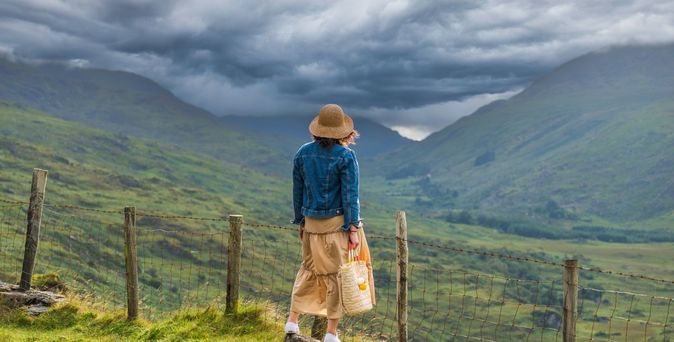 Frau steht auf einem Stein und blickt auf auf eine grüne Berglandschaft